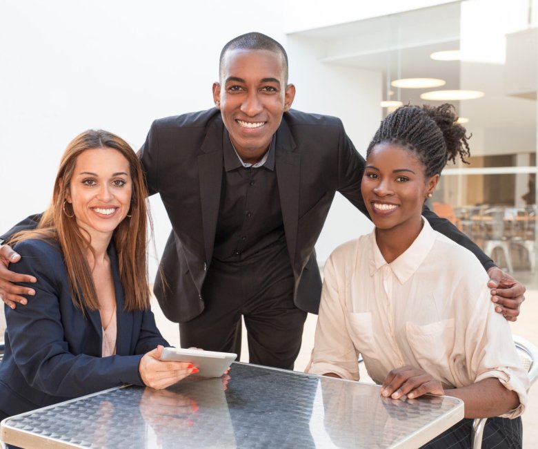 Happy African American businessman embracing female colleagues at cafe. Portrait of three business colleagues sitting at cafe. Successful team concept